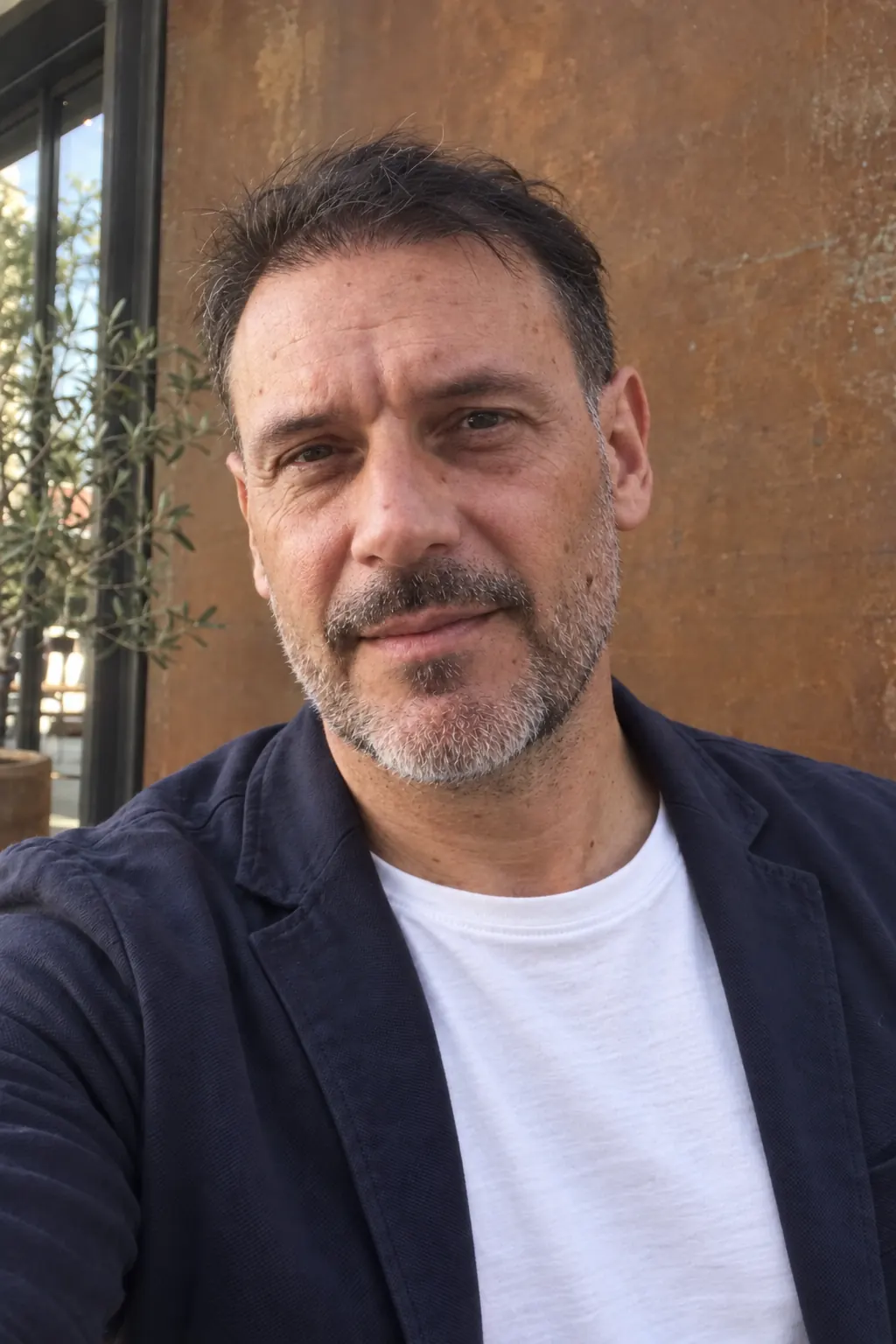 Uomo con capelli scuri e barba, ritratto in primo piano con giacca blu scuro e maglietta bianca