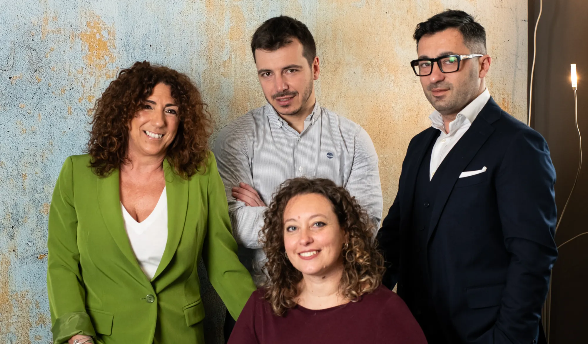Quattro persone sorridono in posa davanti a una parete, tre in piedi e una seduta in primo piano