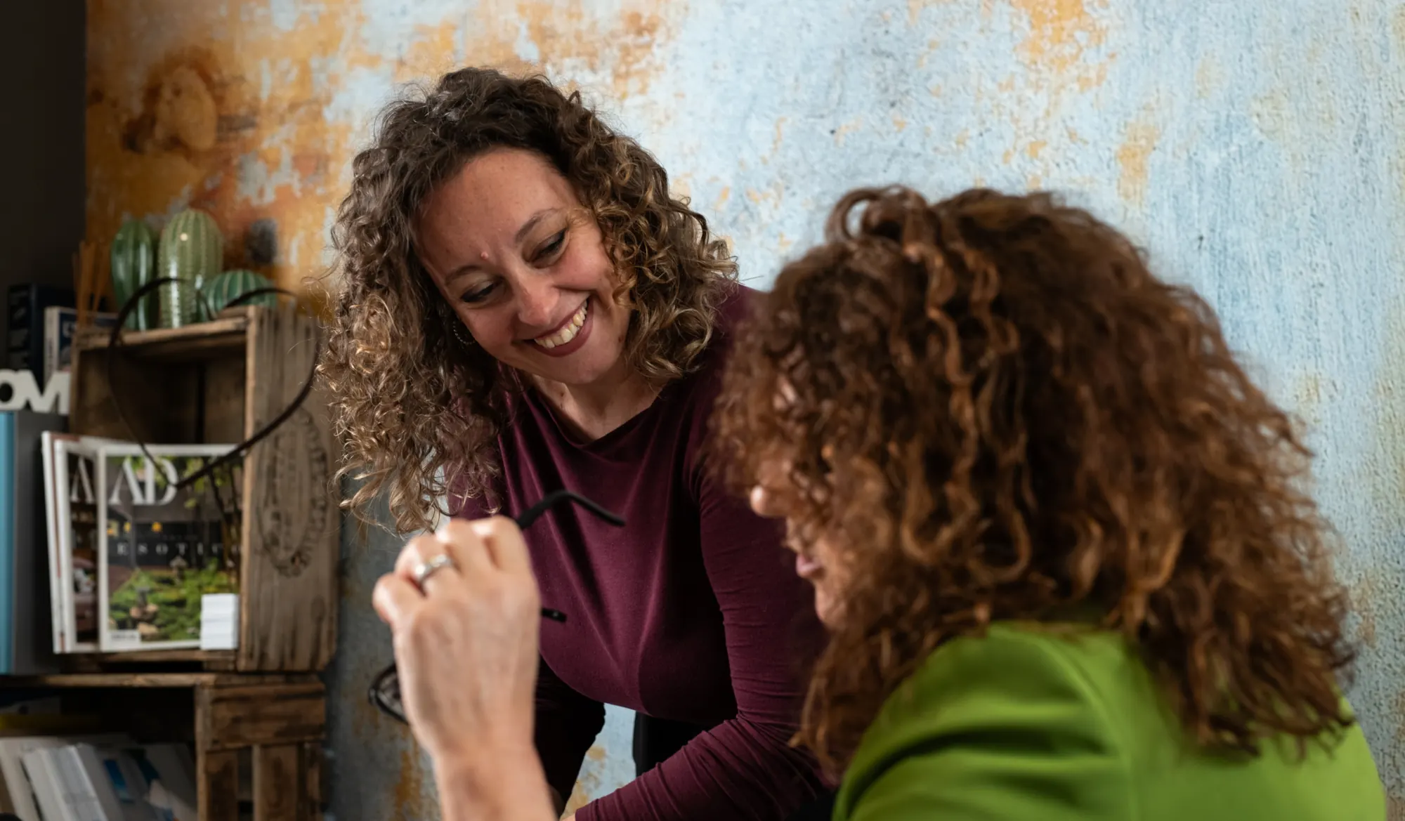 Due donne sorridono durante una conversazione; una tiene in mano un paio di occhiali