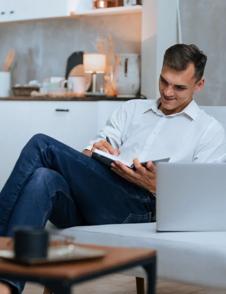 Uomo seduto sul divano che scrive su un taccuino accanto a un laptop