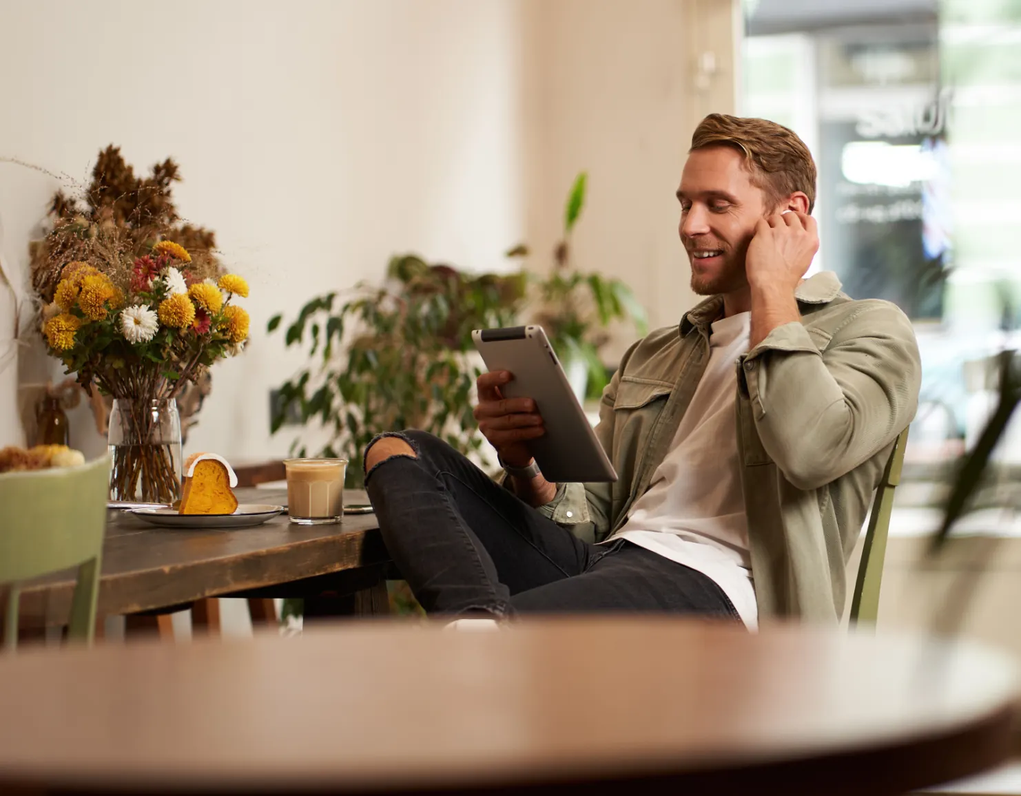 Un uomo sorride mentre usa un tablet al tavolino di un bar, con caffè, fetta di torta e fiori accanto