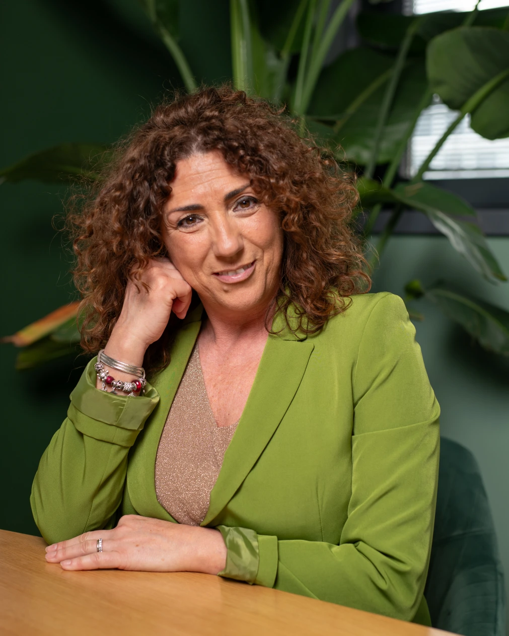 Donna con capelli ricci e blazer verde, seduta a un tavolo e appoggiata con una mano al viso