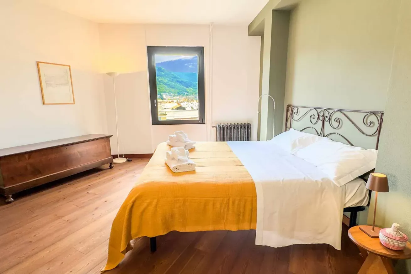 Camera da letto luminosa con letto matrimoniale, coperta gialla, mobili in legno e vista sulle montagne dalla finestra